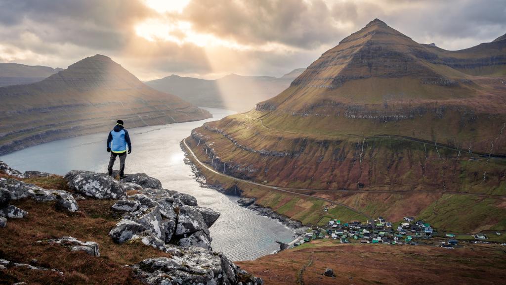 A lone hiker enjoying the panoramic view above Funningur village. The scene captures the scale and beauty of the Faroese fjords on a crisp, clear day.