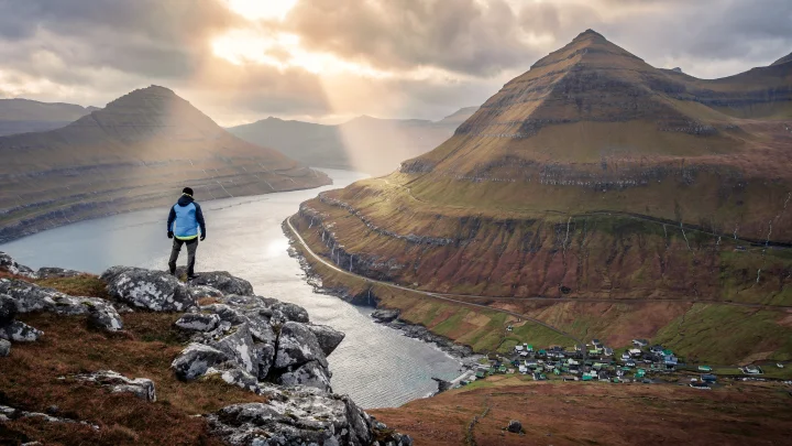 A lone hiker enjoying the panoramic view above Funningur village. The scene captures the scale and beauty of the Faroese fjords on a crisp, clear day.